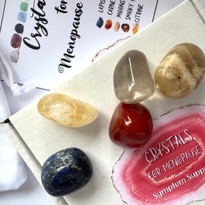 Könnte beinhalten: Ein Set aus sechs Kristallen in einer weißen Box mit einem rosa-weißen Aufkleber, auf dem "Kristalle für die Menopause Symptomentützung" steht. Die Kristalle sind klarer Quarz, Karneol, Citrin, Rauchquarz, Mondstein und Lapislazuli.
