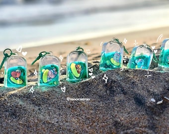 May include: A collection of clear, rectangular keychains with green liquid and cartoon characters. Each keychain has a different character design, with green and teal accents. The keychains are resting on sand near the ocean.