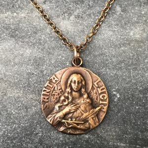 May include: A gold chain necklace with a bronze pendant depicting Santa Lucia. The pendant is oval shaped and inscribed with the words "Santa Lucia" around the edge.