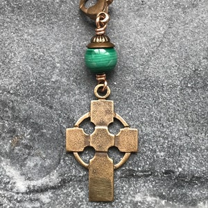 May include: A bronze-toned metal charm featuring a Celtic cross pendant suspended from a green malachite bead and a small bronze-toned lobster clasp. The cross has a circular ring connecting the arms, and the entire piece has a textured finish.