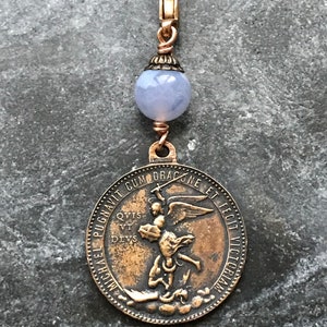 May include: A bronze-coloured charm pendant. It features a round medallion with an image of a figure slaying a dragon, connected to a gold-coloured clasp with a light blue bead.