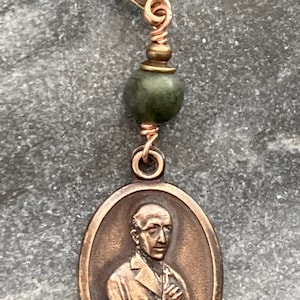 May include: A bronze oval charm with a depiction of a man in a robe with his hands clasped in prayer. The charm is attached to a copper wire with a green stone bead and a gold-toned lobster clasp.