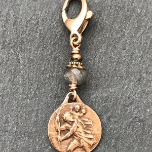 May include: A gold-toned charm with a round, bronze-colored pendant featuring a raised image of a figure holding a spear. The charm is attached to a gold-toned lobster clasp and a gray gemstone bead.
