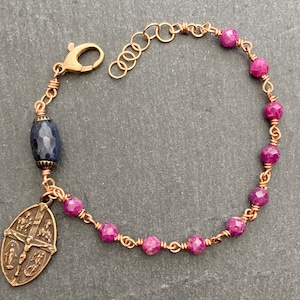 Puede incluir: Un brazalete envuelto en alambre de cobre con una cuenta de piedra azul, un colgante de bronce con un crucifijo y cuatro santos, y cuentas de piedra rosa.