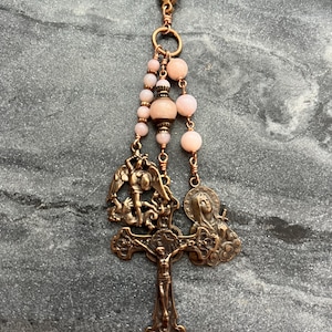May include: A copper-toned metal charm featuring a lobster clasp, three strands of light pink round beads, and three religious pendants. The pendants depict a crucifix, a figure of Saint Michael the Archangel, and a figure of the Virgin Mary.