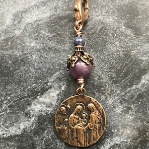 Puede incluir: Un amuleto de bronce con una representación de la Sagrada Familia, con una cuenta de piedra preciosa púrpura y azul y un cierre de bronce.