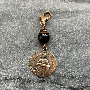 May include: A bronze charm featuring a religious figure with a book and a bird. The charm is attached to a black bead and a gold-toned lobster clasp.