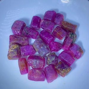 Puede incluir: Colección de piedras preciosas de rubí en bruto en varios tonos de rosa y rojo, con algunos que muestran tonos marrones y dorados. Las piedras en bruto se exhiben en un cuenco blanco, mostrando sus texturas y formas naturales.