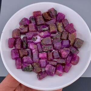 May include: A collection of raw, uncut ruby gemstones in varying shades of pink and red, displayed in a white bowl. The gemstones have a rough, natural texture and are of different sizes and shapes. The bowl is held by a hand.