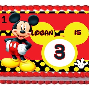 May include: A red, yellow, and black Mickey Mouse themed cake with a white circle with the number 3 in black and red. The text "Logan is 1" is on the cake.