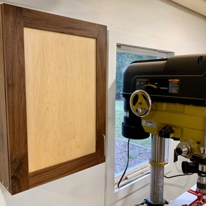 May include: A wooden cabinet with a light wood door and a dark wood frame. The cabinet is mounted on a white wall. A drill press is visible in the background.