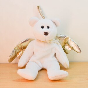 Pode incluir: Um urso de peluche branco com asas douradas. O urso est&aacute; sentado em uma superf&iacute;cie de madeira.