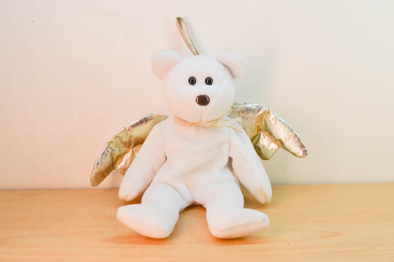 Pode incluir: Um urso de peluche branco com asas douradas. O urso est&aacute; sentado em uma superf&iacute;cie de madeira.