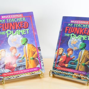 May include: Two copies of the book "My Teacher Flunked the Planet" by Bruce Coville. The book covers feature a green alien, a boy, a girl, and a planet Earth. The books are on gold metal bookends.