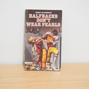 May include: Vintage paperback book titled "Halfbacks Don't Wear Pearls" by Mary Blameslee. The cover illustration depicts two football players in red, yellow, and white uniforms. The book is set against a neutral background.