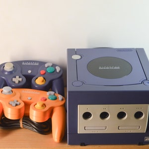 May include: A purple Nintendo GameCube console with two controllers. One controller is purple and the other is orange.