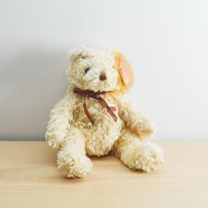 May include: A beige teddy bear with a brown ribbon around its neck. The bear is sitting on a light wood surface.