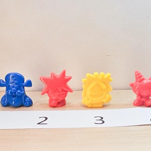 May include: Four small plastic figurines, each with a different design and color. The figurines are numbered 1 through 4. Figurine 1 is blue with a white hat and a white mustache. Figurine 2 is red with a spiky hairdo. Figurine 3 is yellow with a spiky hairdo. Figurine 4 is red with horns and a white face.