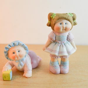 May include: Two porcelain figurines featuring a crawling baby wearing a blue bonnet and a standing child with brown pigtails tied with green ribbons. The baby holds a yellow block with the letter P, while the standing child wears a pastel dress.