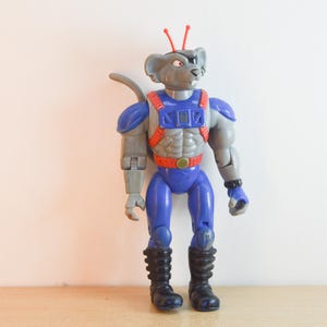 May include: An anthropomorphic mouse action figure with grey skin, wearing a blue and red chest harness, blue trousers, and black boots. The figure has two thin red antennae on its head, a grey tail, and a black eye patch over its left eye.