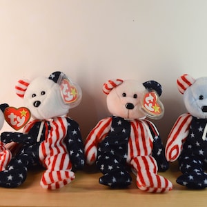 May include: Four plush beanie baby bears wearing red, white and blue patriotic outfits. The bears are sitting on a wooden surface.