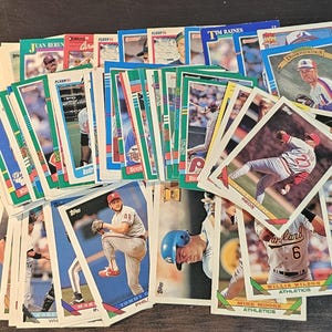 Könnte beinhalten: Eine Sammlung von Baseball-Sammelkarten mit verschiedenen Spielern. Die Karten haben farbenfrohe Designs mit Spielerbildern und Teamlogos. Einige Karten stammen von den Marken Topps und Fleer. Die Karten sind gestapelt und aufgefächert.