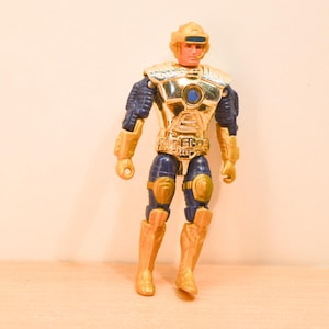 May include: A plastic action figure featuring a gold-colored helmet, chest plate, and limb armor over a dark blue base suit. The figure has a central blue circular detail on the chest, gold gauntlets, knee pads, and boots, with a molded facial expression.