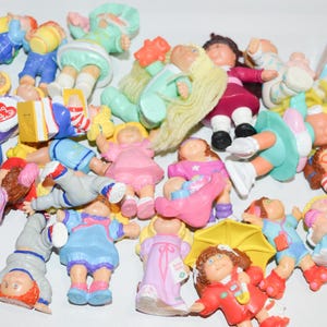 May include: A collection of small, colorful plastic Cabbage Patch Kids figurines in various poses and outfits, including characters with yellow, brown, and red hair, wearing dresses, overalls, and rain gear, some holding accessories like books and umbrellas.