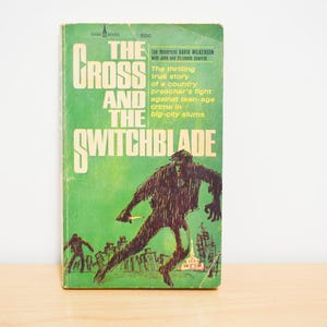May include: Vintage paperback book titled "The Cross and the Switchblade." The cover is green with white lettering and a dark illustration of a figure with a knife. The book's text describes a preacher's fight against crime.
