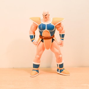 May include: A bald, muscular action figure with a tan complexion, wearing blue and orange armor with shoulder pads, wrist guards, and boots. The figure has a brown belt and a ribbed orange loincloth piece, standing in a forward-facing stance.