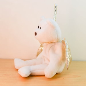 Pode incluir: Urso de pel&uacute;cia branco com asas douradas, sentado em uma superf&iacute;cie de madeira. O urso tem uma aur&eacute;ola e est&aacute; usando um la&ccedil;o branco.