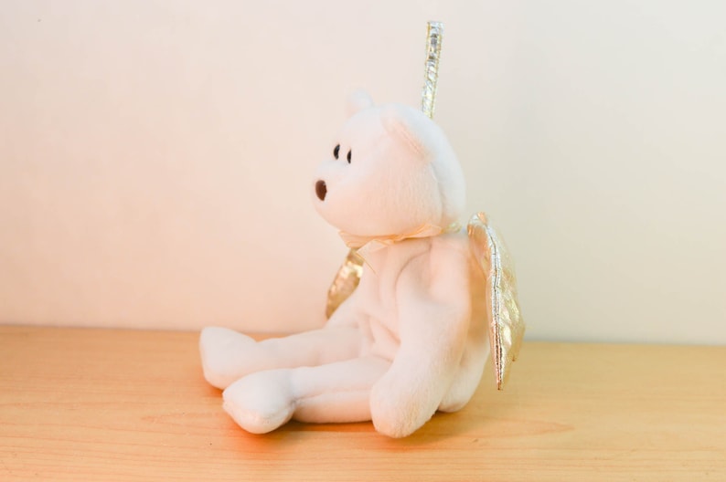 Pode incluir: Urso de pel&uacute;cia branco com asas douradas, sentado em uma superf&iacute;cie de madeira. O urso tem uma aur&eacute;ola e est&aacute; usando um la&ccedil;o branco.