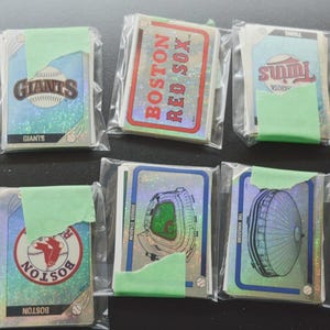 May include: A collection of eight stacks of baseball trading cards, each individually sealed in clear plastic sleeves with light green painter's tape. The cards feature holographic backgrounds and various team logos, stadium illustrations, and team names.
