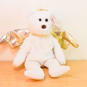 Pode incluir: Urso de pel&uacute;cia branco com asas douradas, usando uma aur&eacute;ola dourada. O urso est&aacute; sentado em uma superf&iacute;cie de madeira.