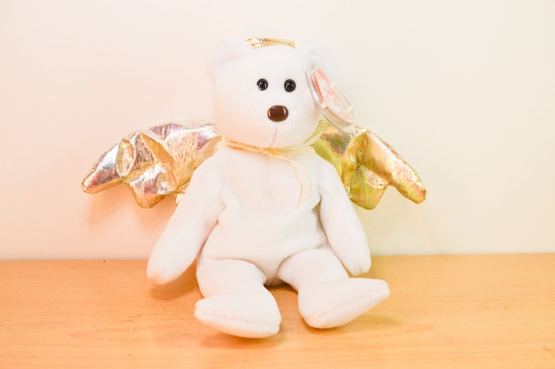 Pode incluir: Urso de pel&uacute;cia branco com asas douradas, usando uma aur&eacute;ola dourada. O urso est&aacute; sentado em uma superf&iacute;cie de madeira.