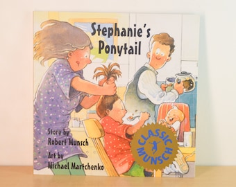 May include: A children's book cover titled "Stephanie's Ponytail" by Robert Munsch with illustrations by Michael Martchenko. The cover features a cartoon illustration of a family in a kitchen. A woman is trying to put a girl's hair in a ponytail. A man is pouring coffee and a baby is sitting in a high chair.