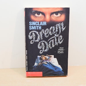 May include: A paperback book titled Dream Date by Sinclair Smith with a black cover. The top features a pair of intense eyes, while the bottom shows a person sleeping in a bed under a blue blanket. The title is written in a stylized diamond font.
