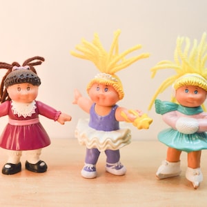 May include: Three small plastic dolls with yarn hair. The first has brown yarn hair and a maroon dress. The second has yellow yarn hair, a purple leotard, and a white tutu. The third has yellow yarn hair, a pink top, a teal skirt, and white skates.