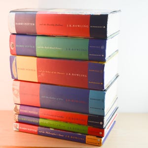 May include: A vertical stack of seven Harry Potter hardback books by J.K. Rowling. The spines are visible, featuring titles including The Deathly Hallows, The Half-Blood Prince, The Order of the Phoenix, The Goblet of Fire, The Prisoner of Azkaban, The Chamber of Secrets, and The Philosopher's Stone, with Bloomsbury and Raincoast logos.