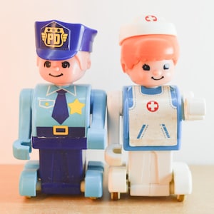 May include: Two plastic toy figures standing side by side. The left figure is a police officer in a blue uniform with a PD cap and yellow star badge. The right figure is a medical professional in a white uniform with a red cross symbol and cap.
