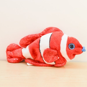 May include: Red and white plush clownfish toy with a blue eye and a blue mouth. The fish is lying on a wooden surface.
