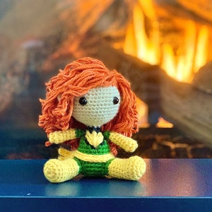 May include: A crocheted doll of a superhero wearing a green and yellow costume with red hair. The doll is sitting on a blue surface in front of a fireplace.
