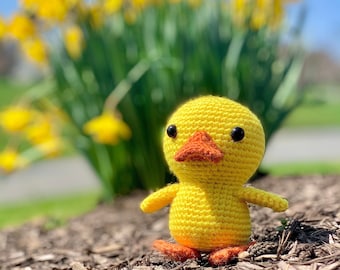 May include: A yellow crocheted duck toy with orange feet and beak, standing on a bed of brown wood chips. The duck is in focus, while a blurred background shows a bed of yellow daffodils.