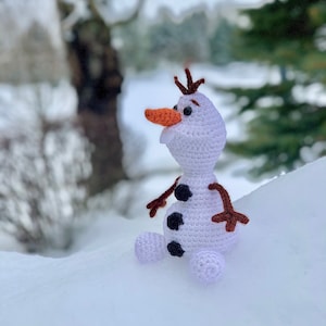 May include: A white crocheted snowman toy with black buttons and an orange nose sitting in the snow.