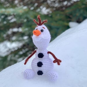 May include: A crocheted Olaf doll from the movie Frozen. The doll is white with a carrot nose, black button eyes, and three black buttons on the body. It has brown twig arms and hair, and is sitting on snow.