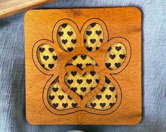 May include: A wooden coaster with a laser-cut design of a paw print with a heart inside. The paw print and heart are filled with a pattern of small black hearts.
