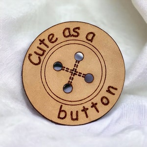 May include: A tan leather button with the text "Cute as a button" engraved around the edge. The button has four holes in the center.