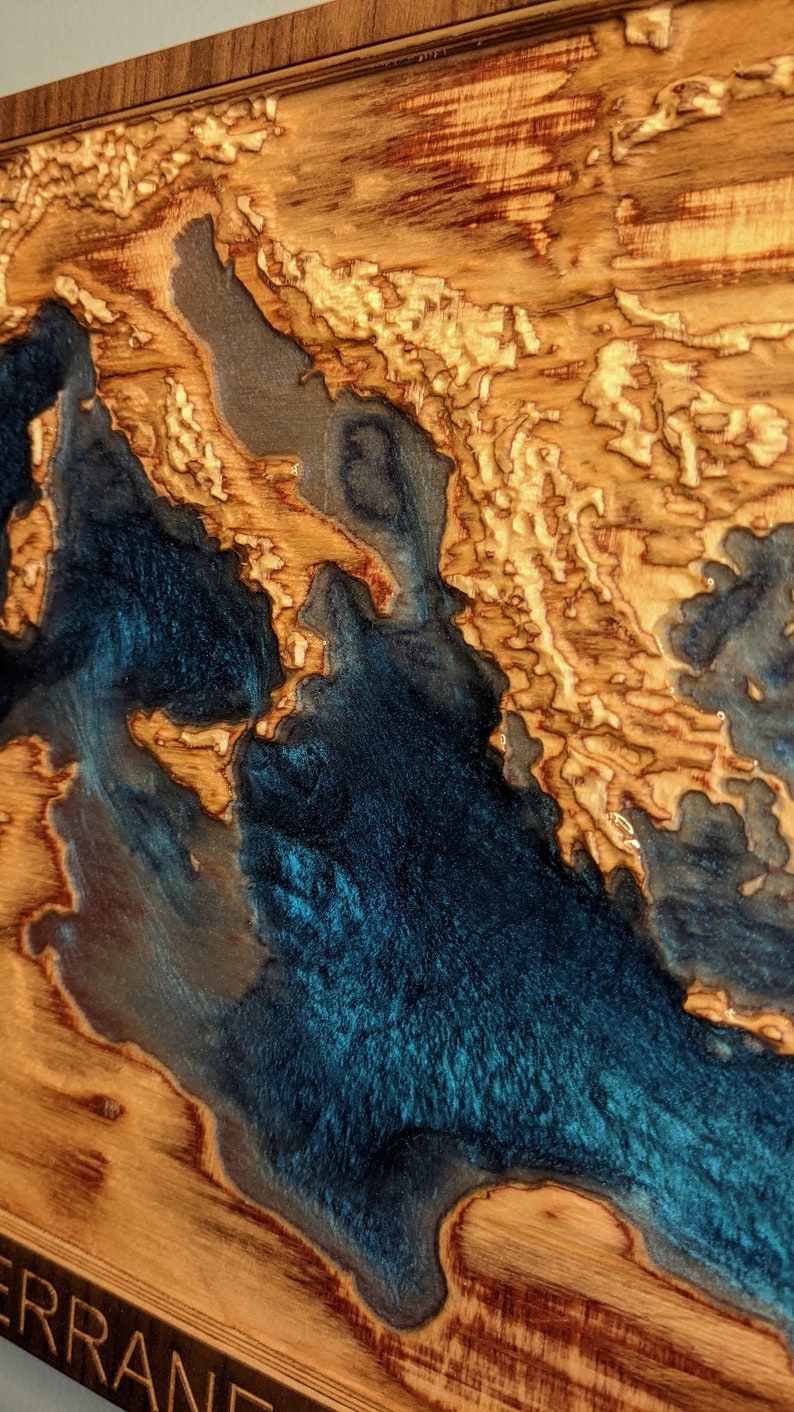K&ouml;nnte beinhalten: Eine Holzkarte des Mittelmeers mit einer blauen Harz-Einlage. Die Karte ist in einem dunklen Holzrahmen gerahmt und tr&auml;gt die Inschrift "MEDITERRANE" am unteren Rand.