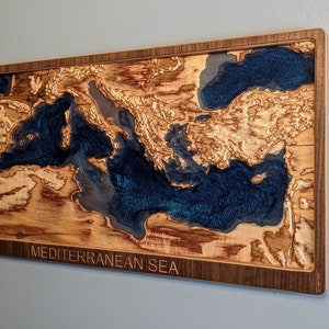 K&ouml;nnte beinhalten: Ein Wandkunstst&uuml;ck aus Holz, das das Mittelmeer darstellt. Das Meer ist mit blauem Harz gef&uuml;llt und das umliegende Land ist aus Holz. Das St&uuml;ck ist in einem dunklen Holzrahmen gerahmt und tr&auml;gt die Worte "MEDITERRANEAN SEA" unten eingraviert.