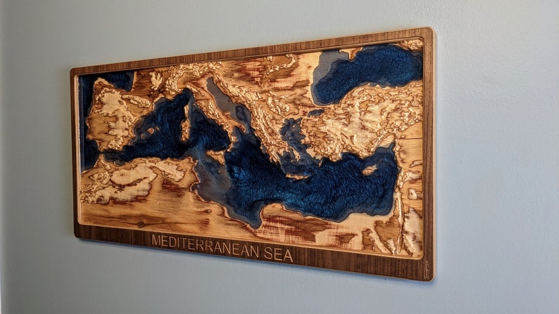 K&ouml;nnte beinhalten: Ein Wandkunstst&uuml;ck aus Holz, das das Mittelmeer darstellt. Das Meer ist mit blauem Harz gef&uuml;llt und das umliegende Land ist aus Holz. Das St&uuml;ck ist in einem dunklen Holzrahmen gerahmt und tr&auml;gt die Worte "MEDITERRANEAN SEA" unten eingraviert.